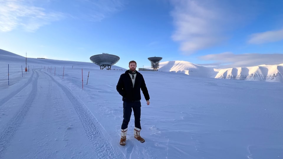 junger Mann mit rötlichem Haar und Bart, dick eingepackt auf Schnee, hinter ihm Schneelandschaft.