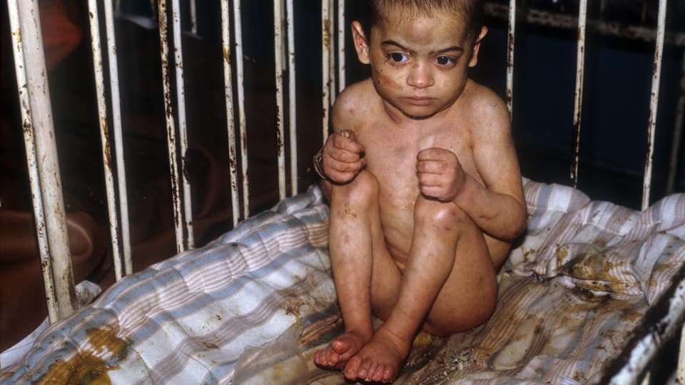 Ein verkrustetes Kleinkind sitzt nackt auf einer kotbeschmutzten Matraze in einem rostigen Bett mit hohen Gittern.