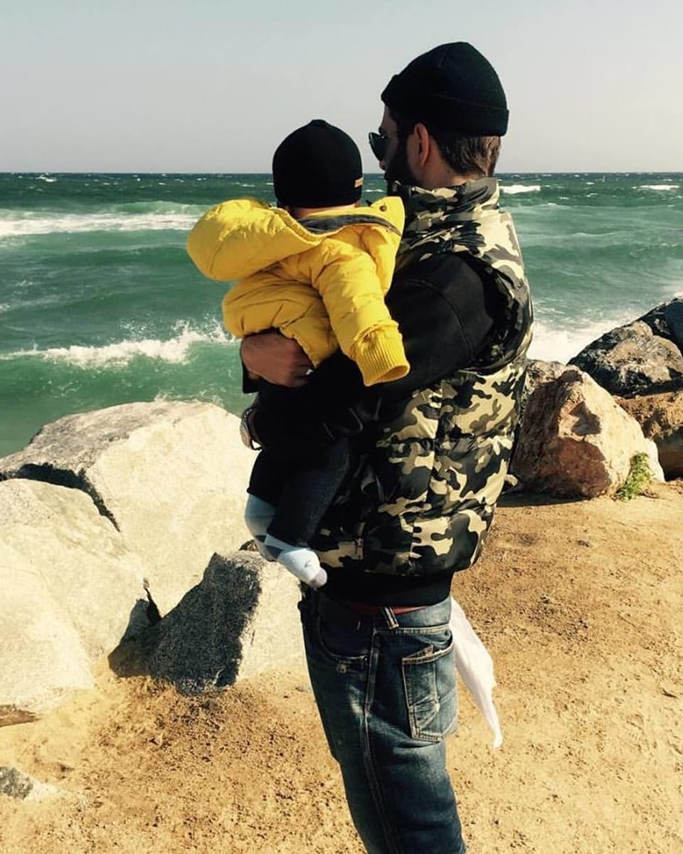 Bligg steht mit seinem Sohn im Arm vor dem Meer. Beide mit dem Rücken zu uns gewandt.