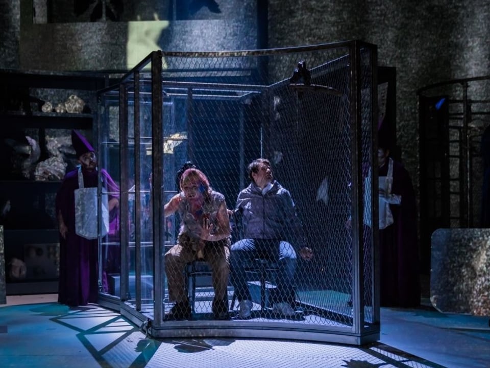 Zwei Männer sitzen in einem grossen Käfig auf einer Bühne.