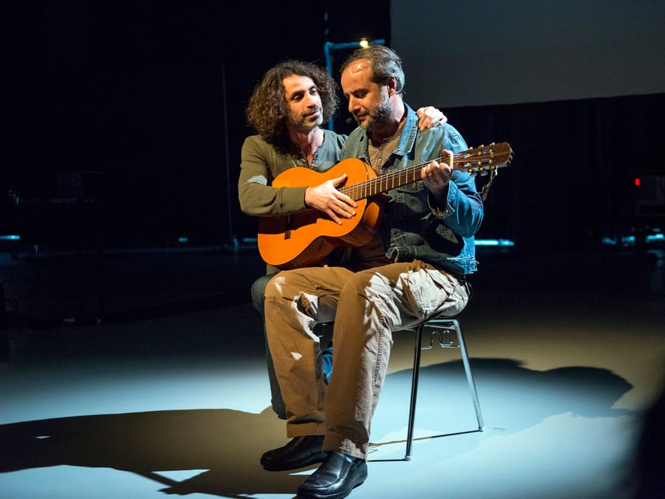 Ein Mann sitzt mit Gitarre auf der Bühne, ein anderer Mann legt den Arm um ihn und redet ihm zu.