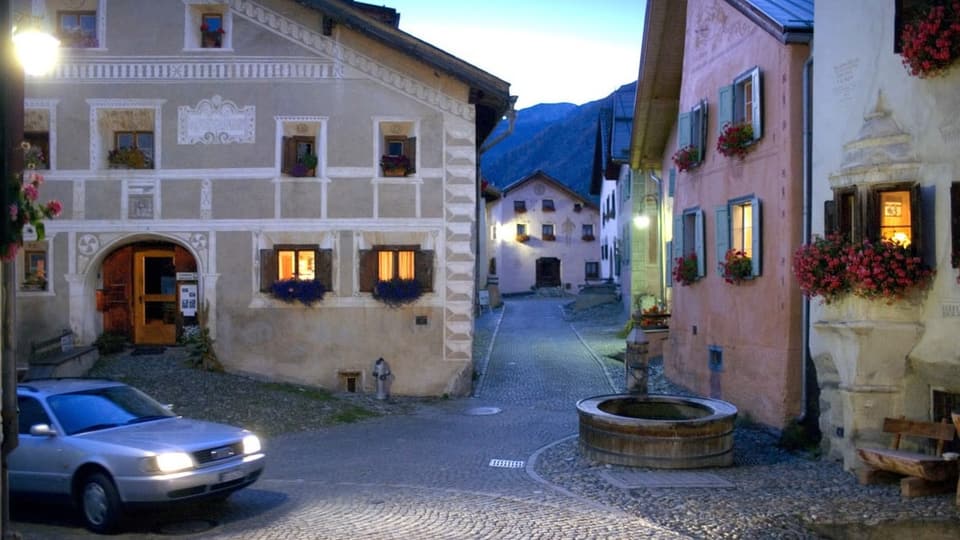 Dorfplatz mit alten Häusern, Dorfbrunnen und Auto auf der Strasse. 