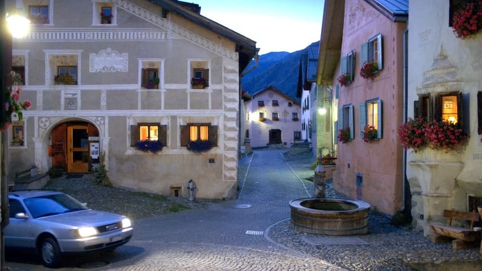 Dorfplatz mit alten Häusern, Dorfbrunnen und Auto auf der Strasse.