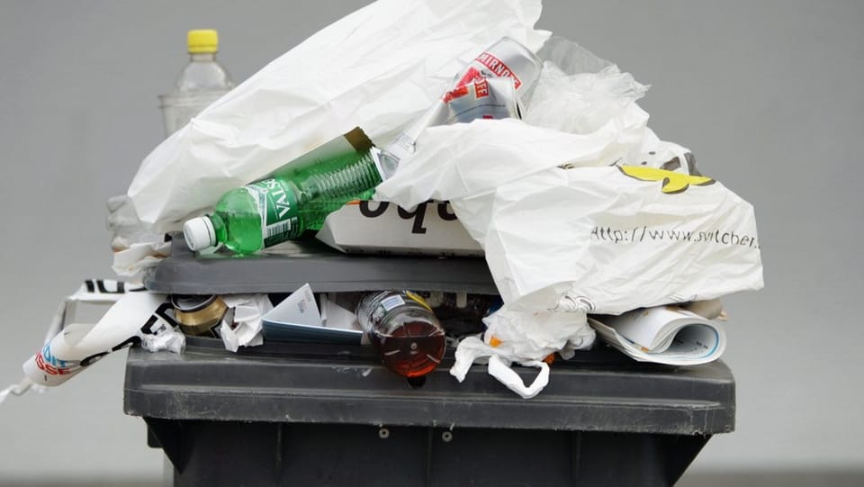 Eine schwarze überfülte Abfalltonne. Plastiksäcke und PET-Flaschen fallen heraus.