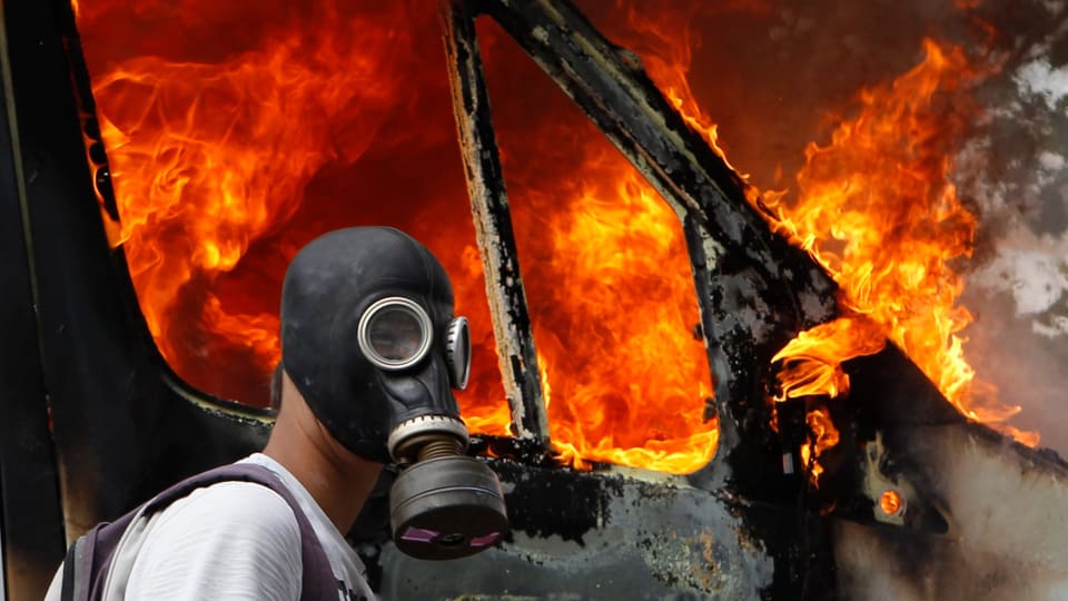 Symbolbild: Person mit Gasmaske vor brennendem Auto.