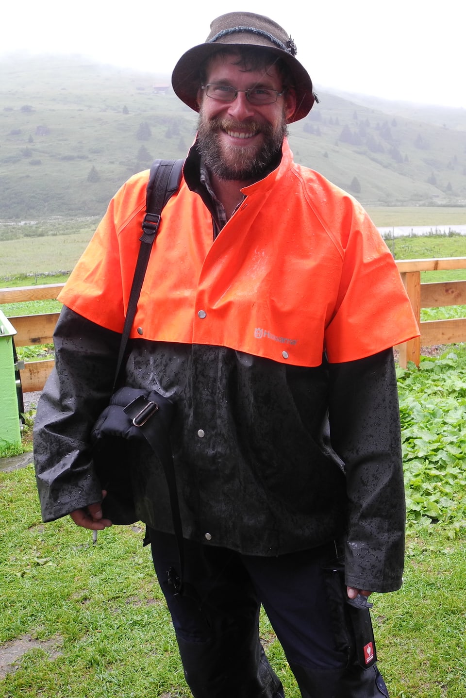 Ludwig Willmann mit orangefarbenem, regendichtem Umhang über wetterfester Kleidung und seiner Tasche mit wichtigen Utensilien über der Schulter.