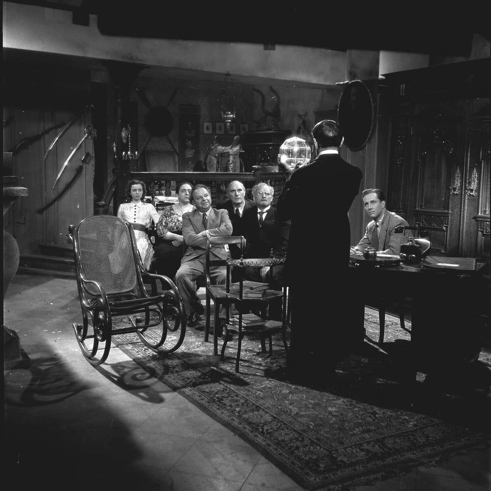 Sechs Männer und zwei Frauen sitzen in einem kleinen Saal. Einer der Männer hält eine Ansprache.