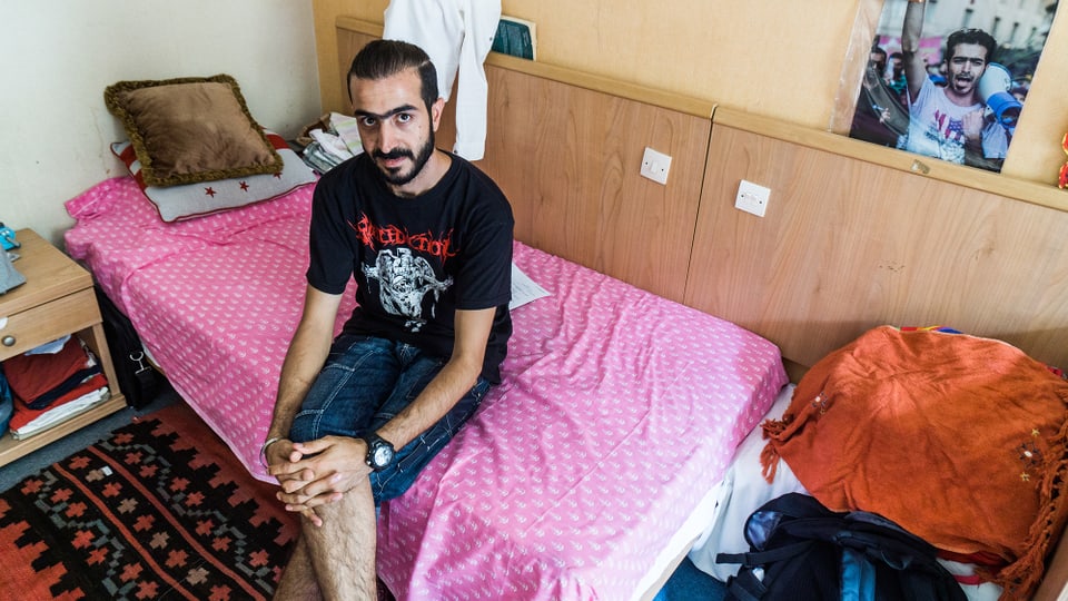 Ein Mann sitzt mit übereinandergeschlagenen Beinen auf dem Bett eines Hotelzimmers.