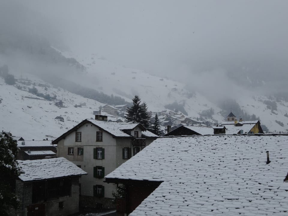 Im Dorf liegt Schnee auf den Hausdächern. Im Hintergrund die frisch verschneiten Berghänge.