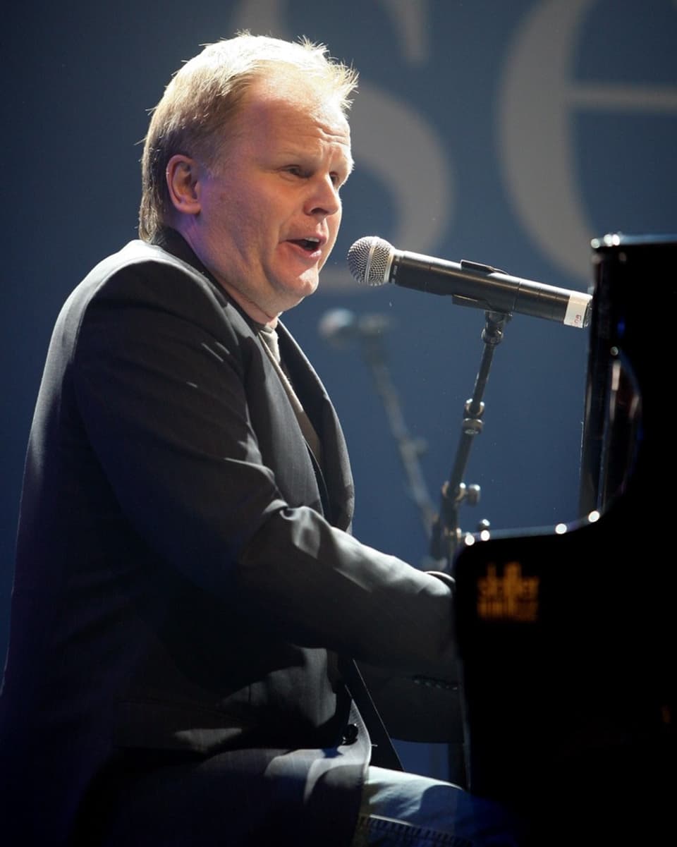 mittelalter Mann sitzt am Klavier, seitlich fotografiert, in dunkelgrauem Blazer, singt in Mikrofon.