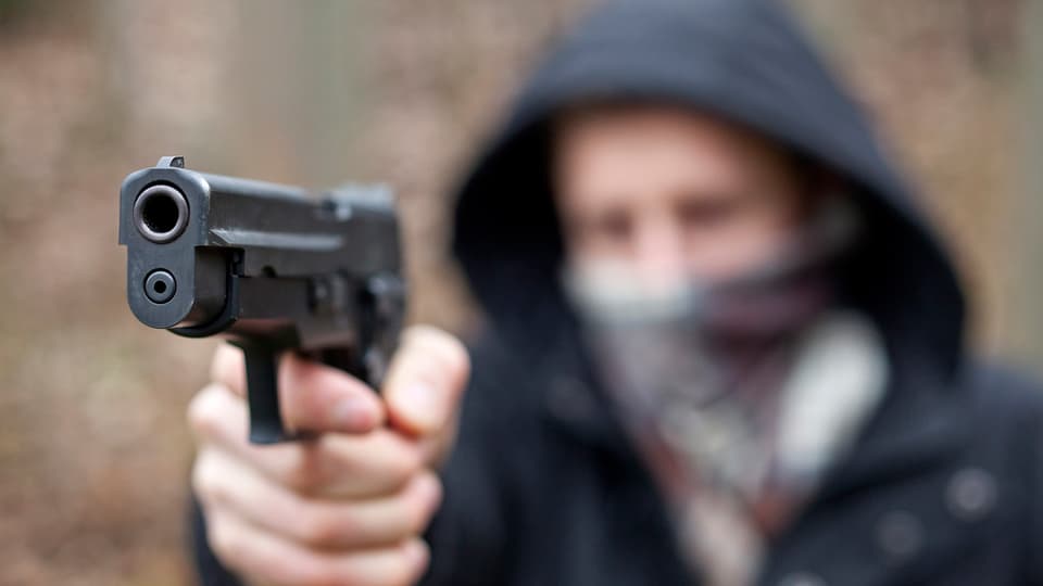 Verschwommene Aufnahme von einem maskierten Mann mit einer Pistole im Vordergrund.