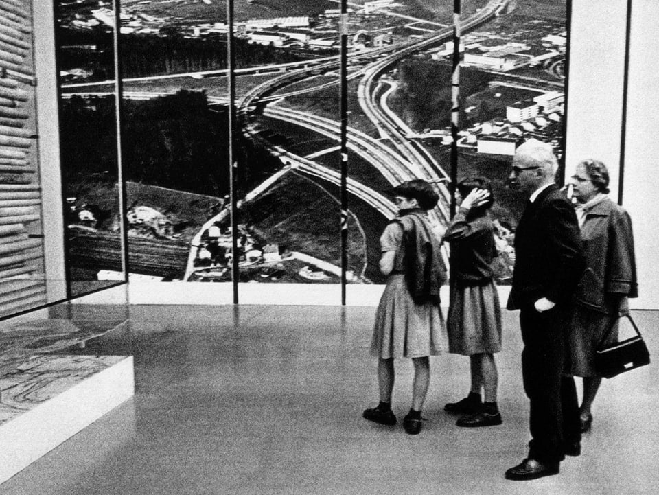 Auf einer schwarz-weissen Fotografie stehen ein paar Menschen vor einem grossen Bild, auf dem eine Autobahn-Schlaufe zu sehen ist.