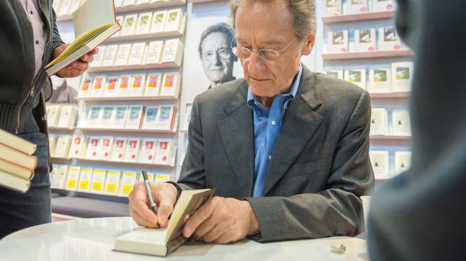Ein alter Mann im Anzug und mit Brille sitzt an einem Tisch und signiert ein Buch. Hinter ihm Regale mit Büchern