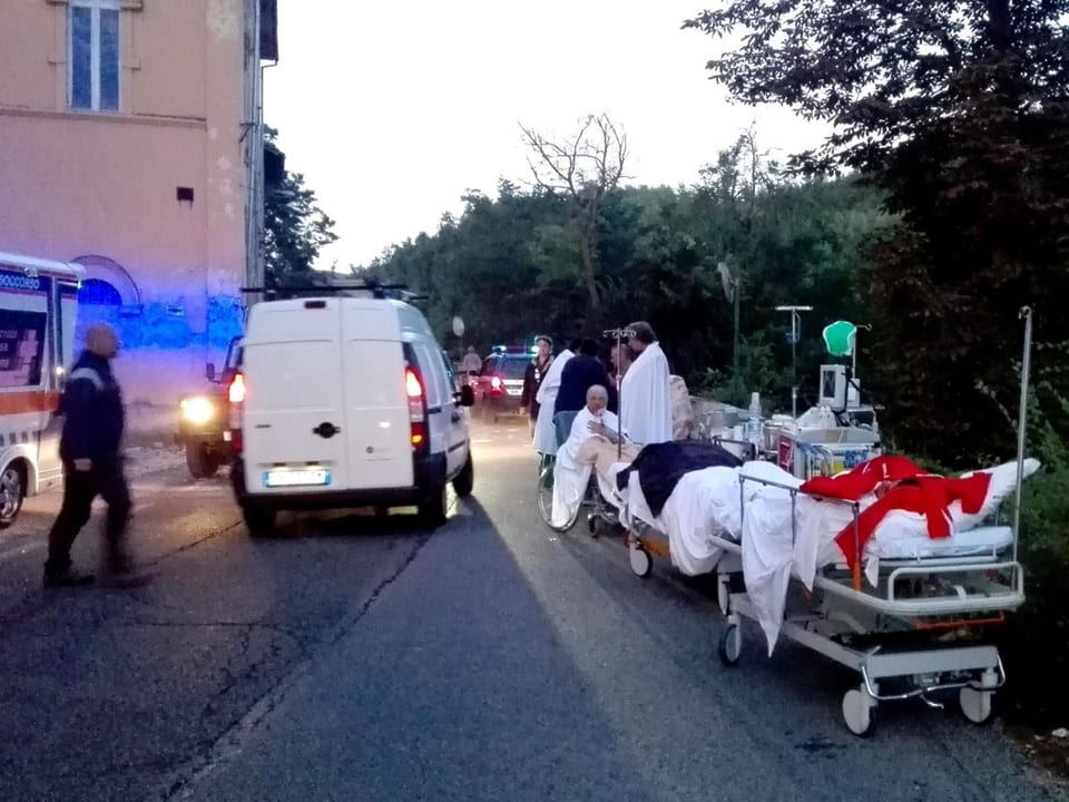 Krankenwagen und Bahre stehen auf einer Strasse, darum herum Patienten