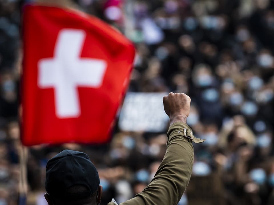 Ein Mann erhebt bei einer Demo die Hand zur Faust, dahinter schwingt die Schweizer-Flagge.