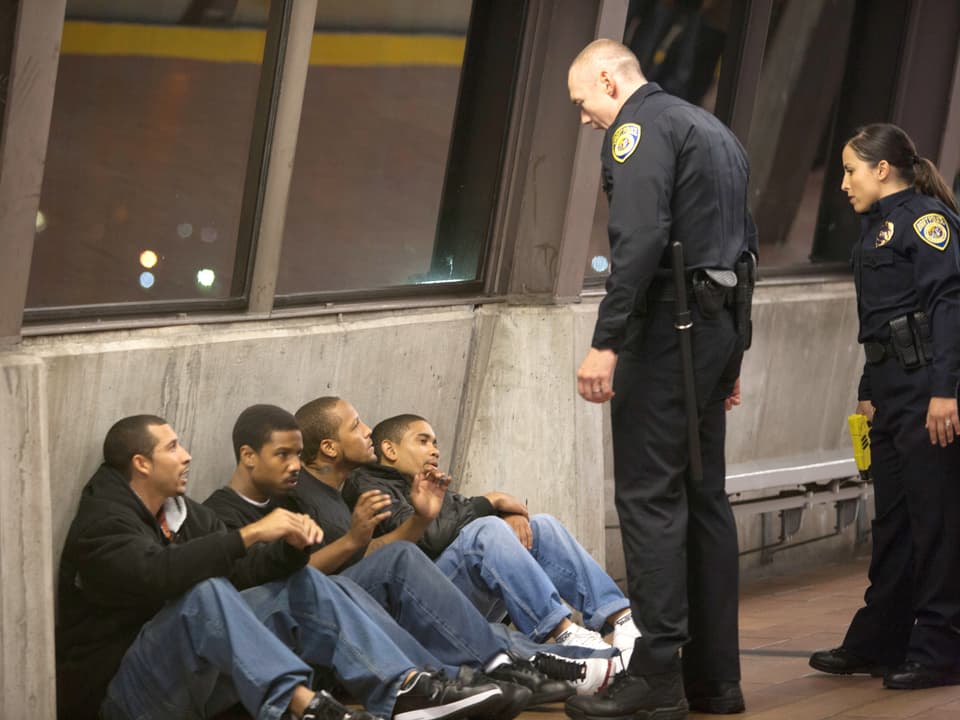 Vier junge Männer sitzen mit dem Rücken zu einer Betonwand, ein Polizist und eine Polizistin stehten vor ihnen.