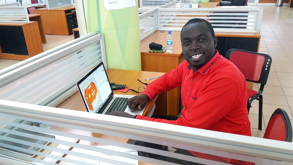 Ein Mann sitzt lächelnd an einem Schreibtisch mit Laptop.