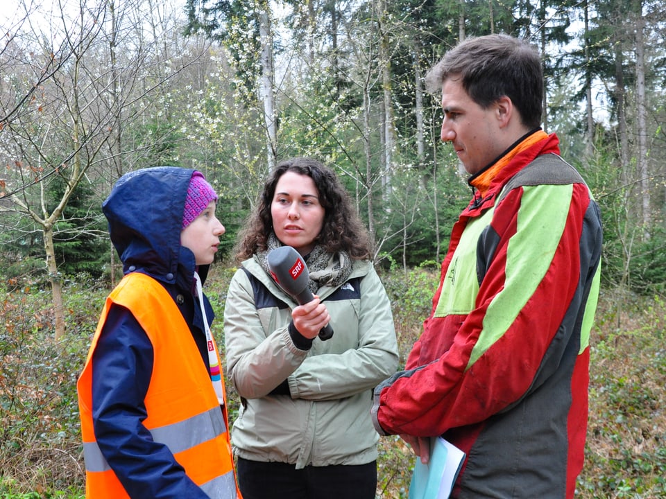 Ein Mädchen, eine Reporterin und ein Forstwart stehen im Wald
