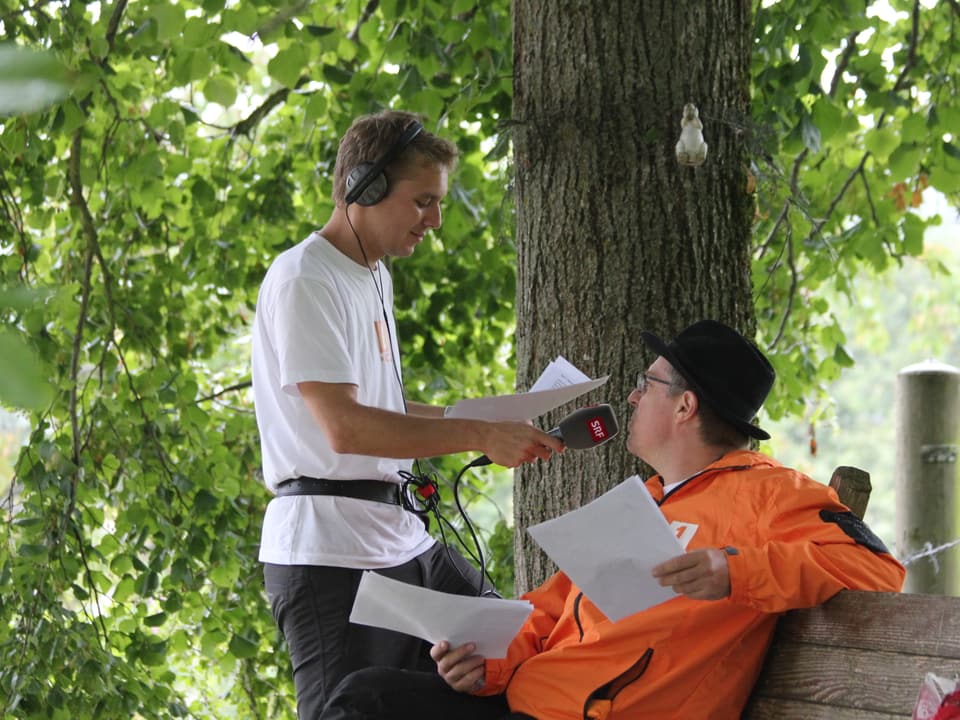 Reto Scherrer interviewt einen Kollegen der auf einer Bank unter einem Baum sitzt.