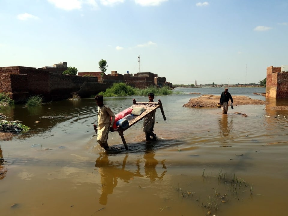Zwei Männer tragen im knietiefen Wasser ein Möbelstück. Die Umgebung mit Häusern ist überschwemmt.