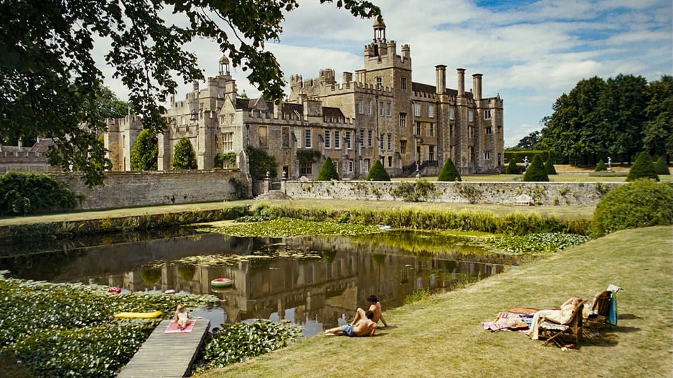 Auf dem Bild ist ein Schloss zu sehen, eine grosser Rasen, Liegestühle und drei Personen.
