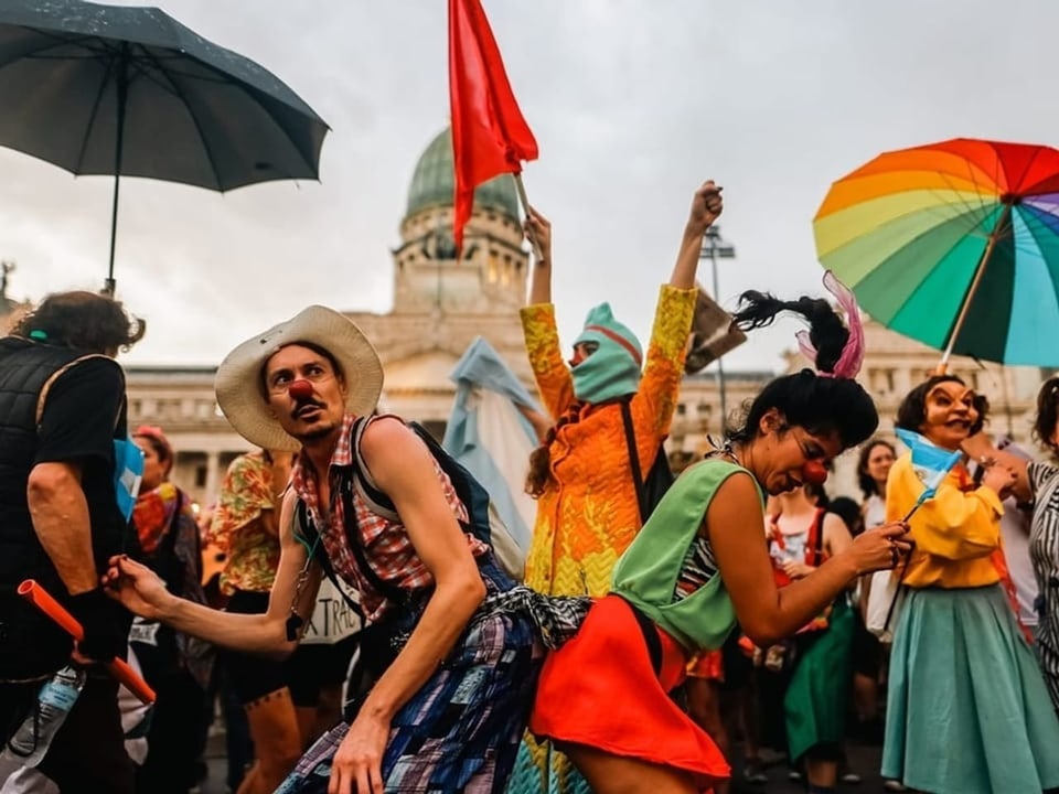 People wearing colorful clothes and holding rainbow-colored umbrellas dance in front of the National Congress.