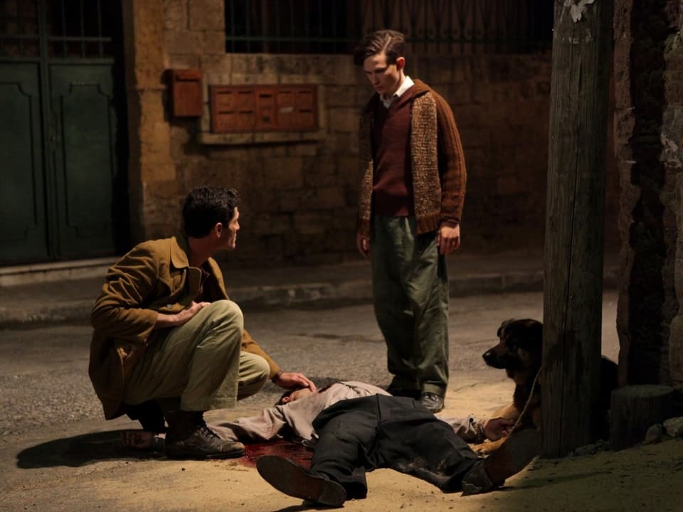 Zwei Männer, einer kniend, der andere stehend, neben einer Leiche.