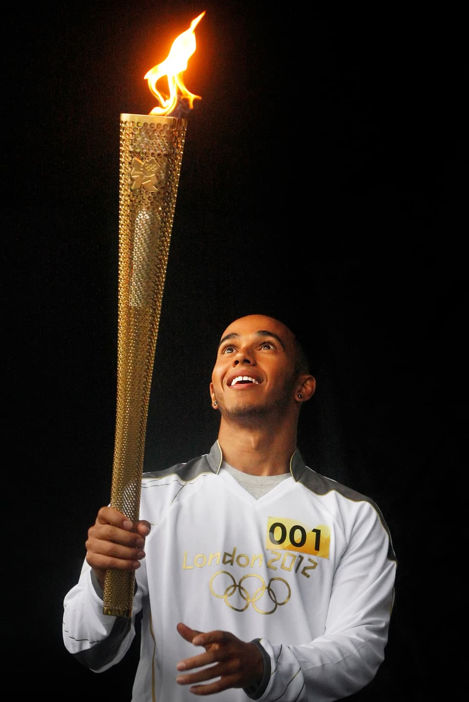Lewis Hamilton trägt mit einem breiten Grinsen die Olympische Flamme.
