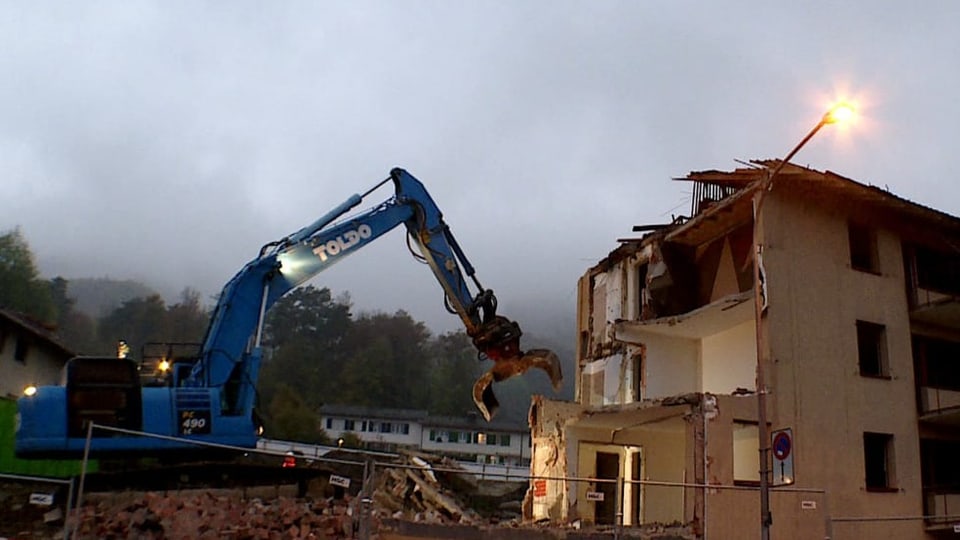 Ein Bagger vor einem halb zertrümmerten Wohnhaus im Abendrot.