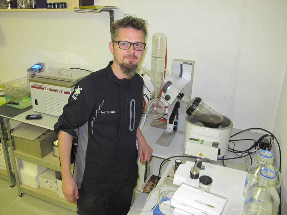 Mann mit Brille und Spitzbart in schwarzen Kleidern mit Aufdruck "Rolf Caviezel" zwischen Labor-Geräten