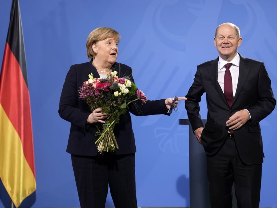 Angela Merkel (links) und Olaf Scholz stehen nebeneinander auf einer Bühne. Merkel hält einen Blumenstrauss.