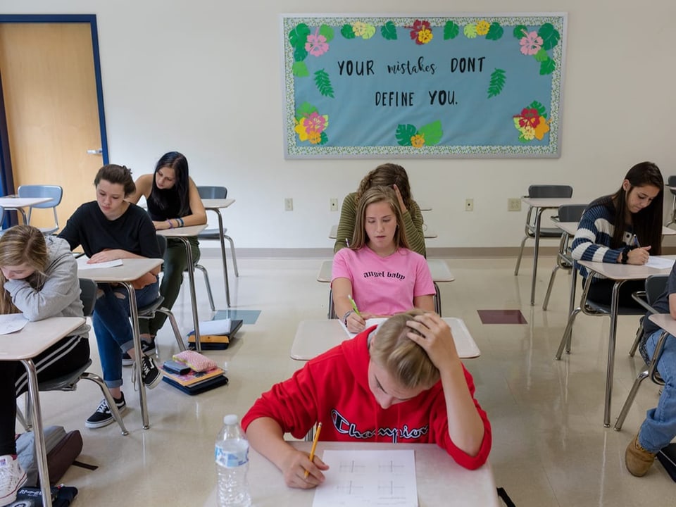 Foto Klassenzimmer, drei Tischreihen. In der Mitte Mädchen mit langen, dunkelblonden Haaren und pinkem T-Shirt.