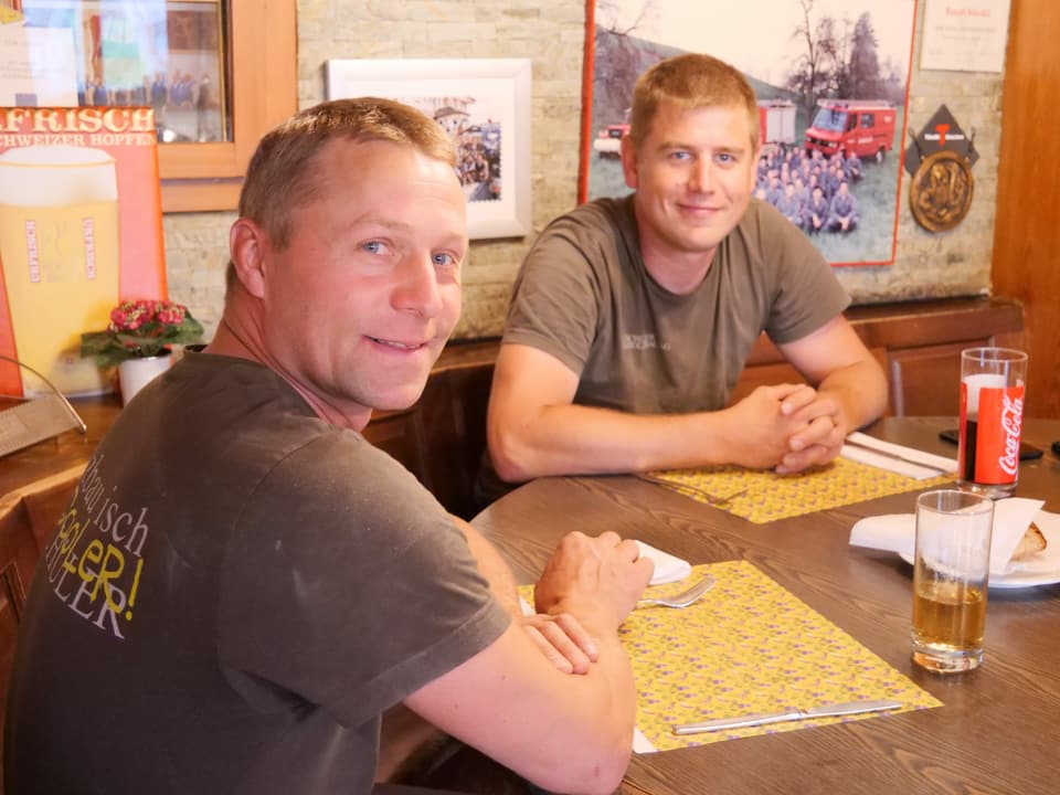 Zwei Arbeiter in braunen T-Shirts sitzen am Tisch. Vor sich ein Papierset und Süssgetränke. 