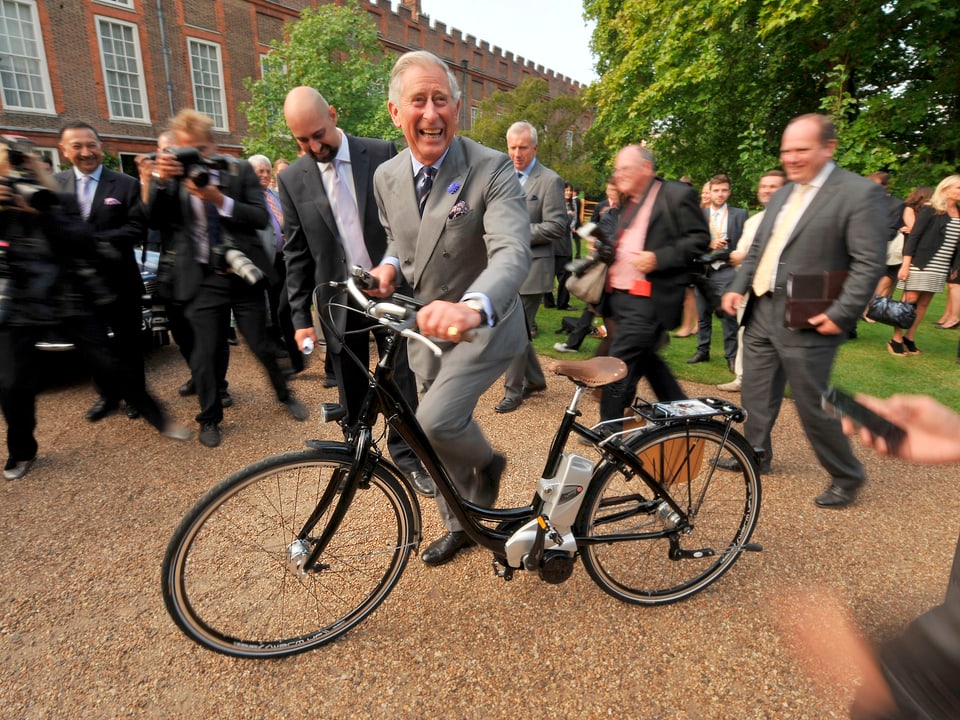 Prinz Charles in einem grauen Anzug auf ein schwarzes Fahrrad sitzend.