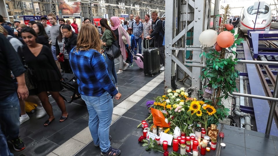 Bahnhofhalle mit Geleisen. Daneben eine Gedenkstätte mit Blumen und Trauerkerzen.