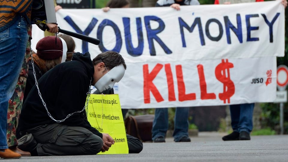 Ein Mann mit Maske kauert vor einem Plakat mit Aufschrift «your money kills»
