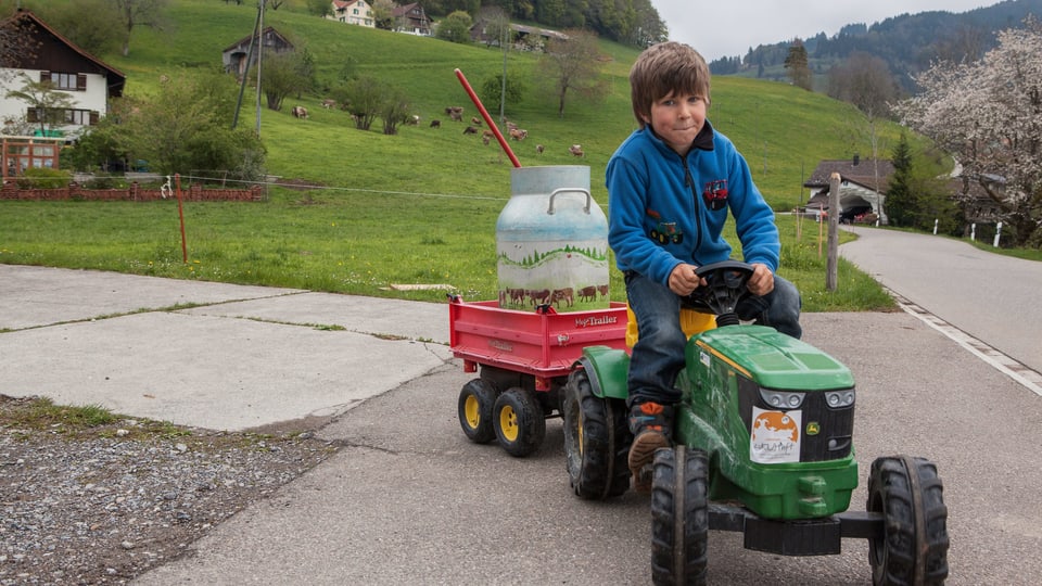 Ein Kind fährt mit einem Spielzeugtraktor. Auf einem Anhänger steht eine Milchkanne.
