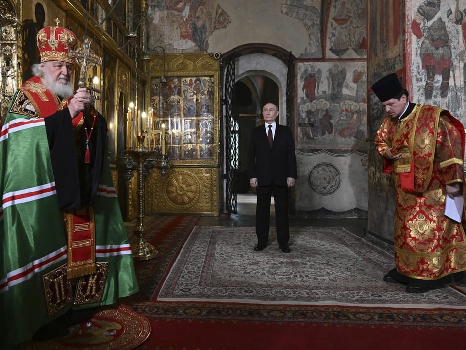 Putin in einer Kathedrale. Links hält ein Geistlicher ein Kreuz in der Hand. Rechts verneigt sich ein Geistlicher