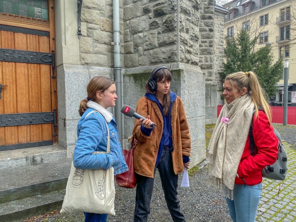 Ein Mädchen und zwei Frauen stehen vor einer Kirche am Stauffacher. Das Mädchen spricht ins  Mikrofon.