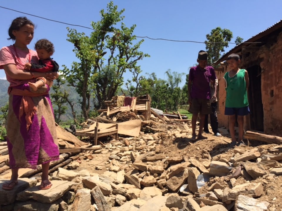 Eine Frau mit Kleinkind im Arm steht vor den Trümmern eines Hauses, daneben zwei Jungen. 