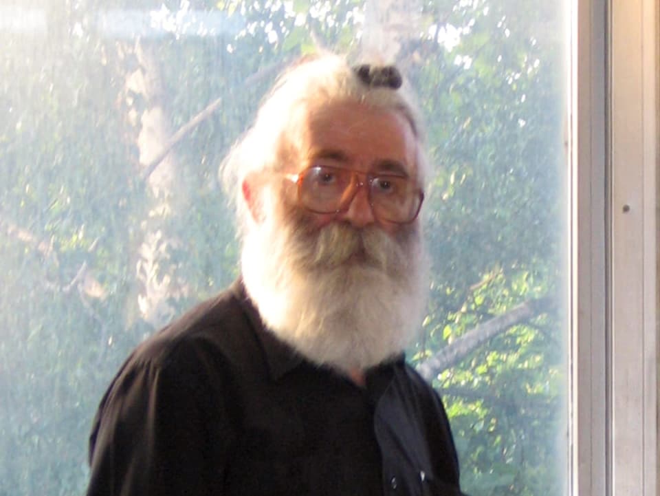 Radovan Karadzic steht mit langem wiessen Bart und Brille vor einem Fenster. 