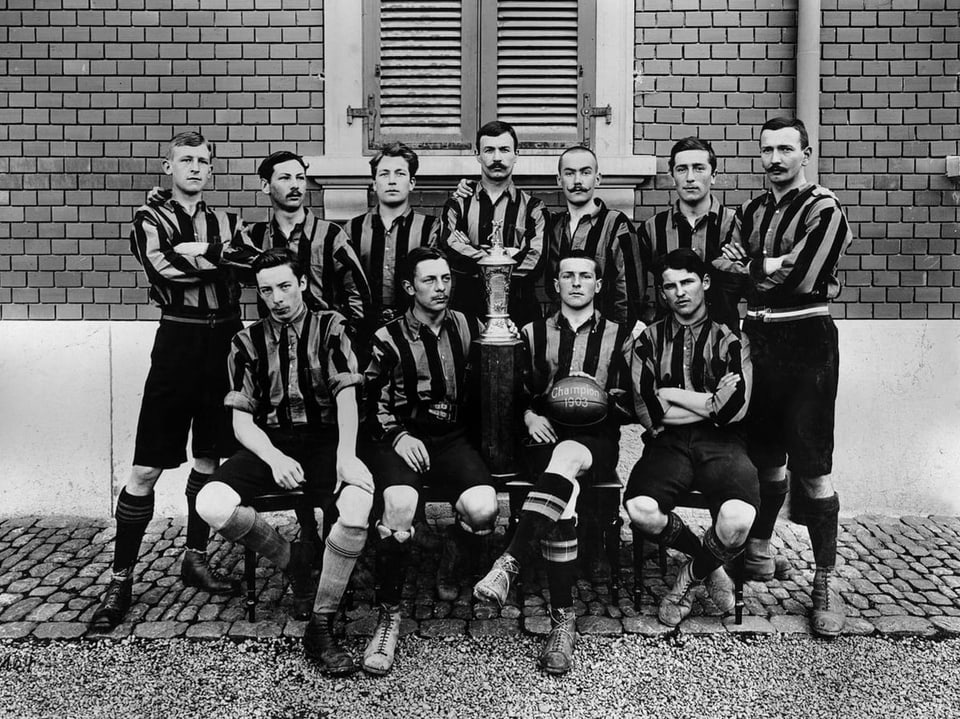 Schwarzweiss-Bild mit elf Männern in gestreiften Trikots, die um einen Pokal versammelt sind.