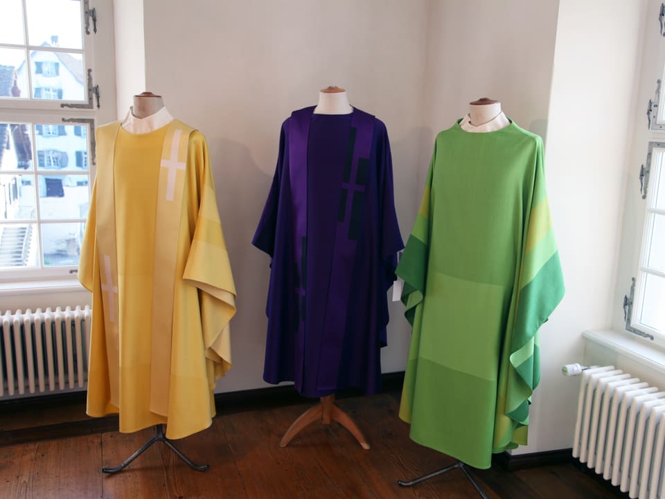 Auf dem Bild sind drei liturgische Gewänder ausgestellt. Ein Gelbes, ein Violettes sowie ein Grünes. 