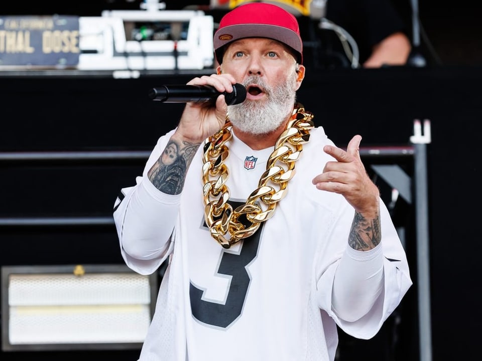 Mann mit rotem Hut und goldener Kette singt in ein Mikrofon auf einer Bühne.