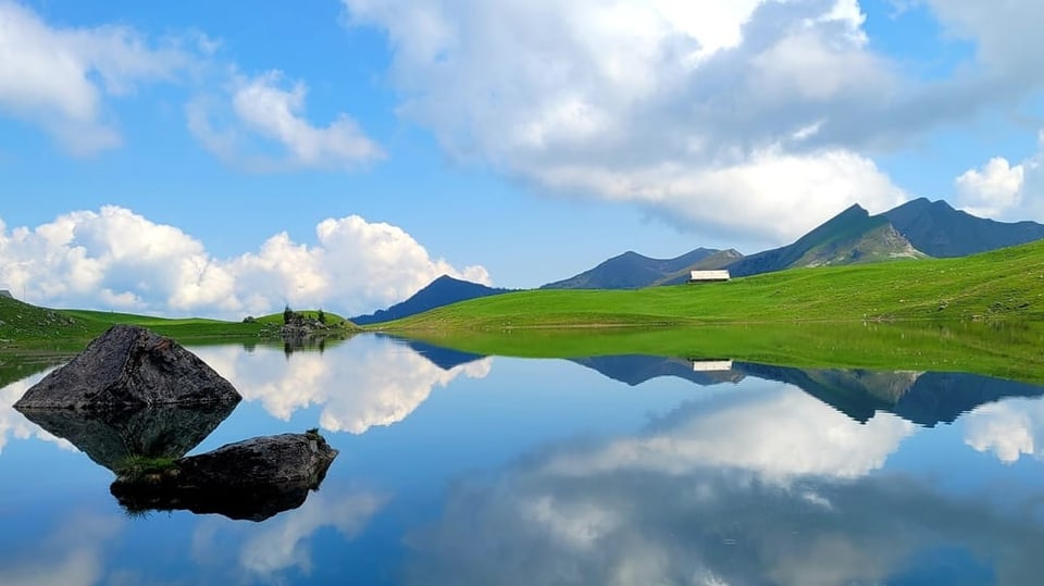 Grüne Alp mit See, in dem sich blauer Himmel mit Quellwolken spiegeln. 