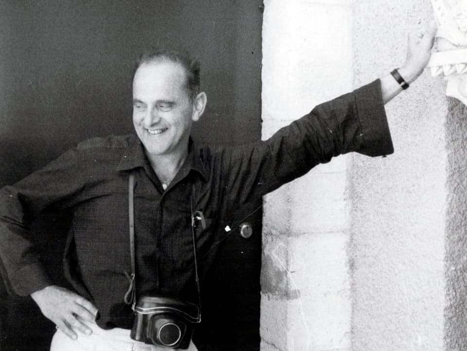 Schwarzweissfoto: Mann mit Analogkamera lehnt sich lächelnd an Säule