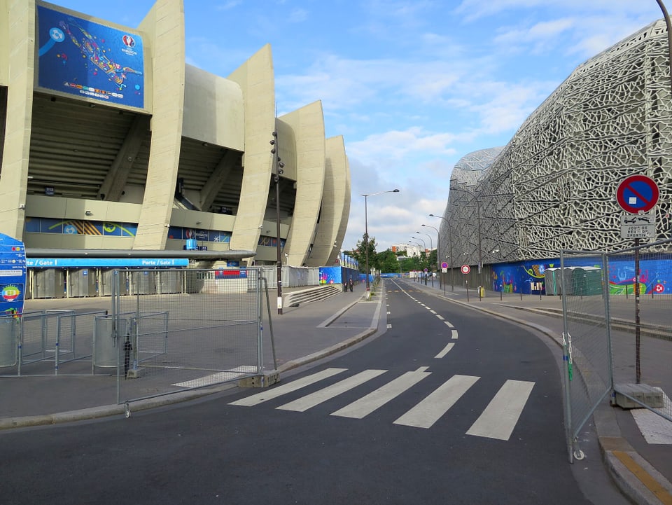 Strassenaufnahme. Auf der linken Seite steht ein Stadion, rechts ist ein Gebäude zu sehen.
