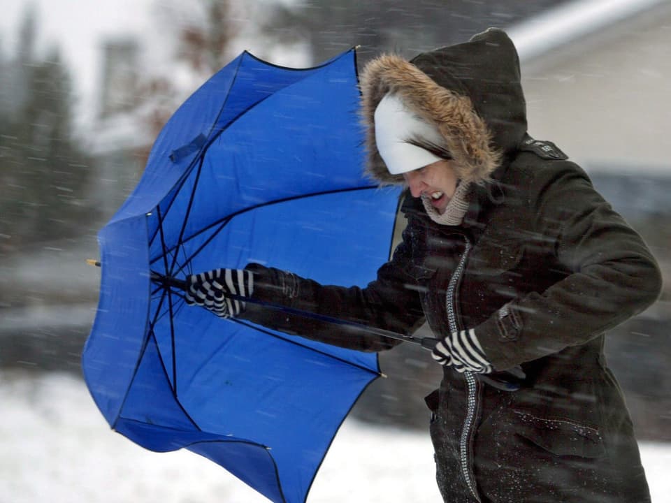 Der Sturm fegt Schnee  durch die Ortschaft, ein Frau kämpft mit geöffneten Schirm dagegen an.