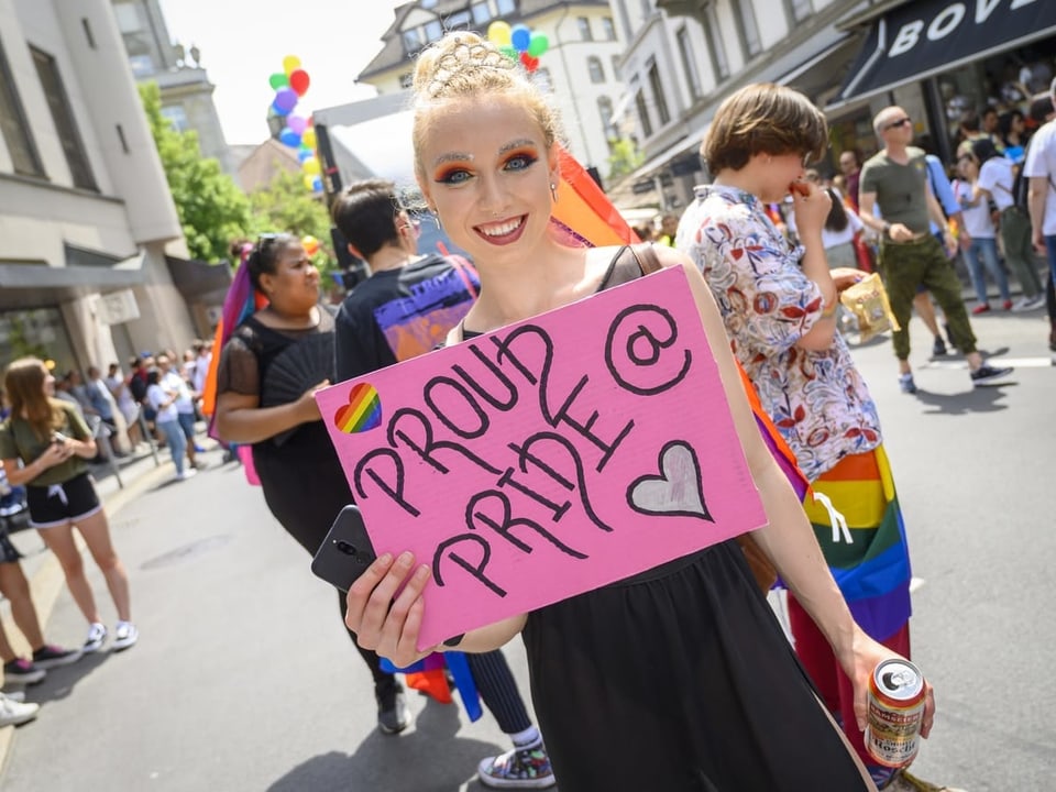 Eine junge Frau mit blonden Haaren hält ein pinkes Schild in die Höhe auf dem steht "Proud @ Pride". 