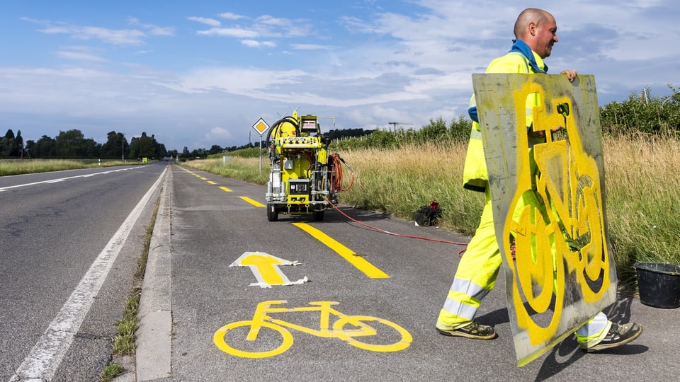 Ein Mann in gelber Leuchtkleidung trägt eine Schablone. Am Boden ist ein gelbes Fahrrad Piktogramm.
